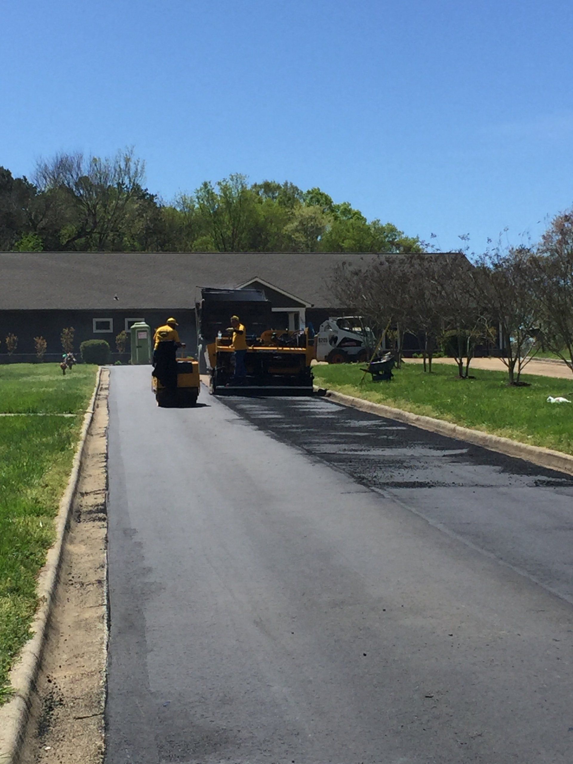 Asphalt paving of a road with machinery and workers under a blue sky, near a building.