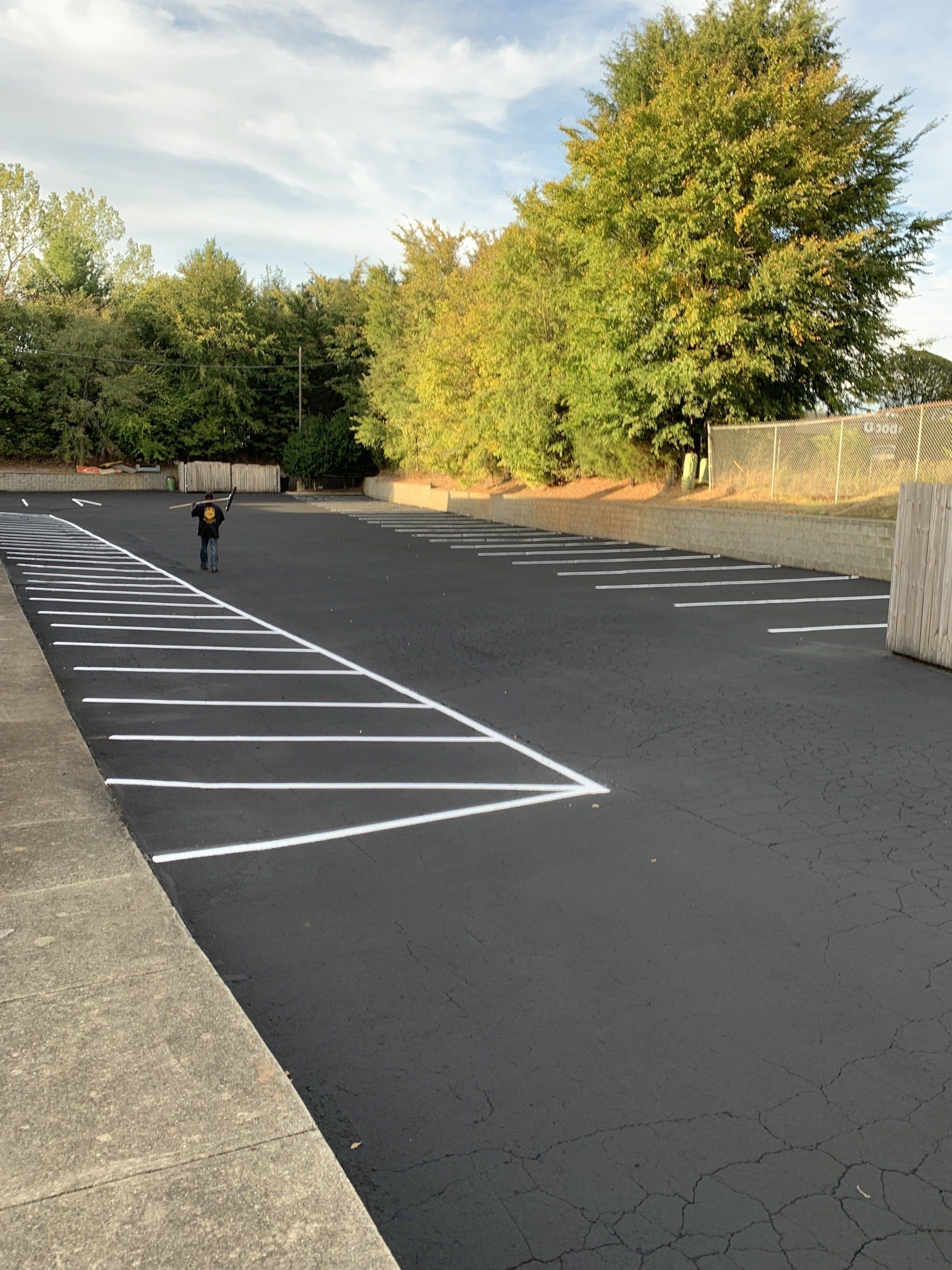 Newly paved parking lot with white painted parking space lines. A person walks in the distance. Trees in the background.