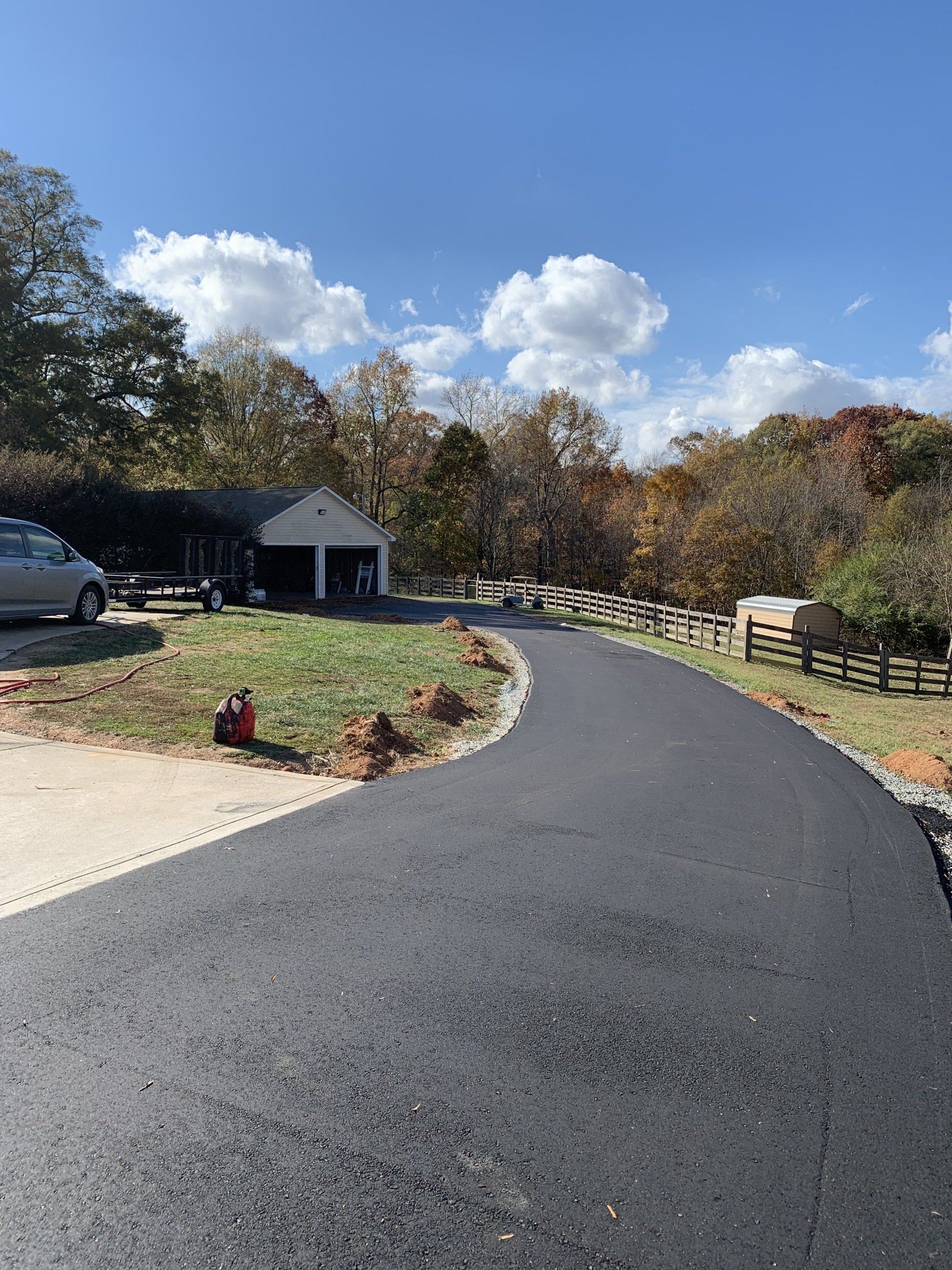 Black asphalt driveway curves toward a house with a garage, trees, and a partly cloudy sky.