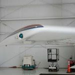 White aircraft wing in a hangar, with equipment beneath; bright light shining from the wing.