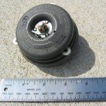Circular black object with metal components, next to a ruler.