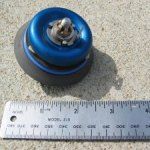 Blue and black object with a nut and ruler for scale on concrete.