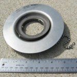 Metal toilet flange with chain, next to a ruler, on a cement surface.