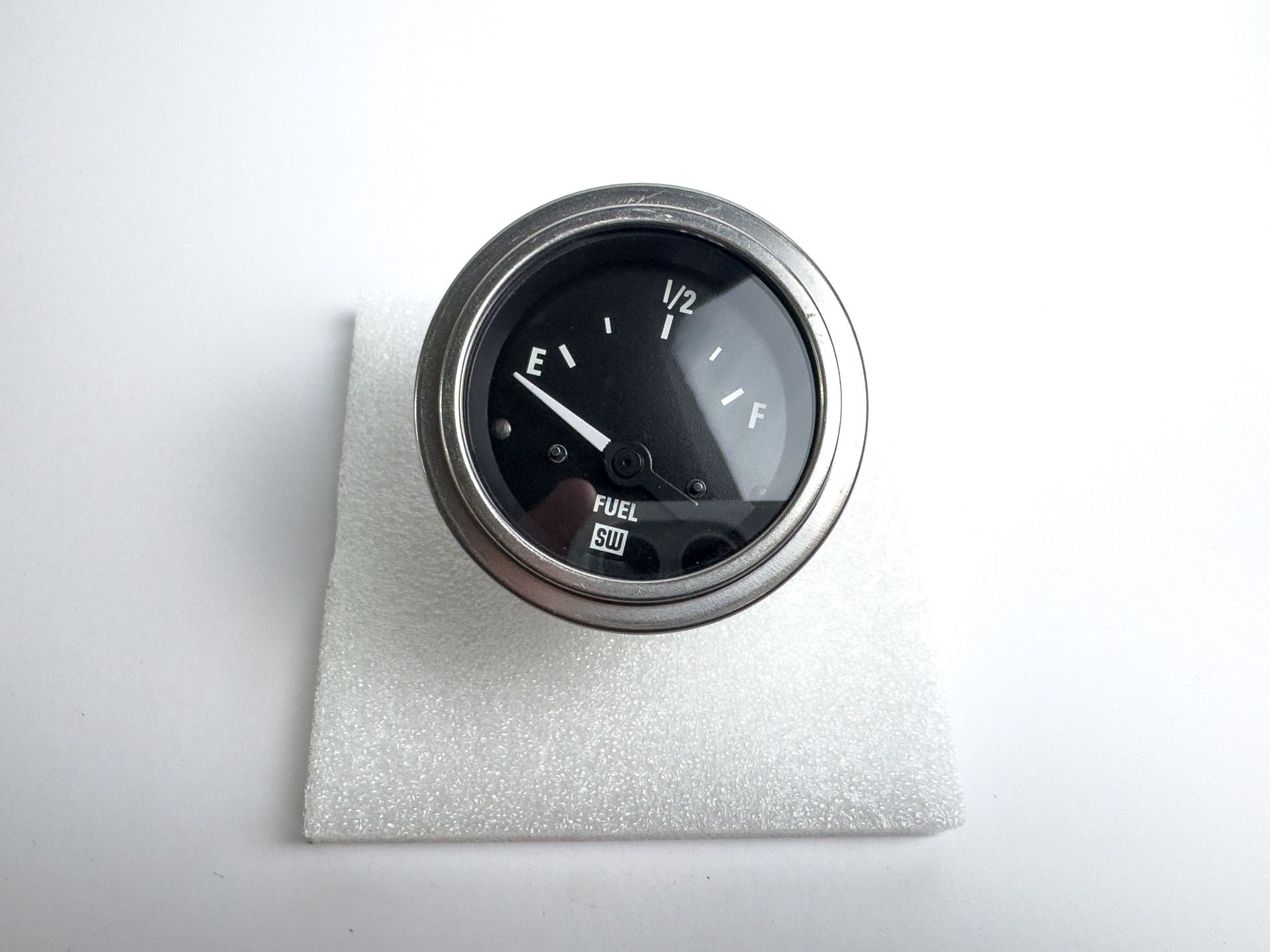 Fuel gauge showing below half full, in a metal bezel, sitting on white foam.