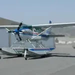 Seaplane on floats, parked on tarmac. Mostly silver with blue and red accents. Propeller in front.