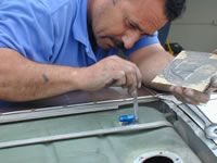 Mechanic working on a fuel tank, using a tool.  Blue shirt, focused expression, shop setting.