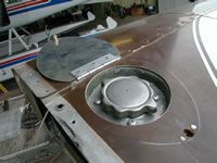 Aircraft wing with open fuel cap and access cover. Brown metal with white markings, aircraft in background.