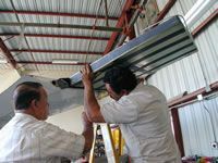 Two men install aircraft component in a hangar. One stands on a ladder, supporting the part.
