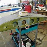 Wing of a plane on a workbench in a workshop; grey metal, green paint, and several circular openings.