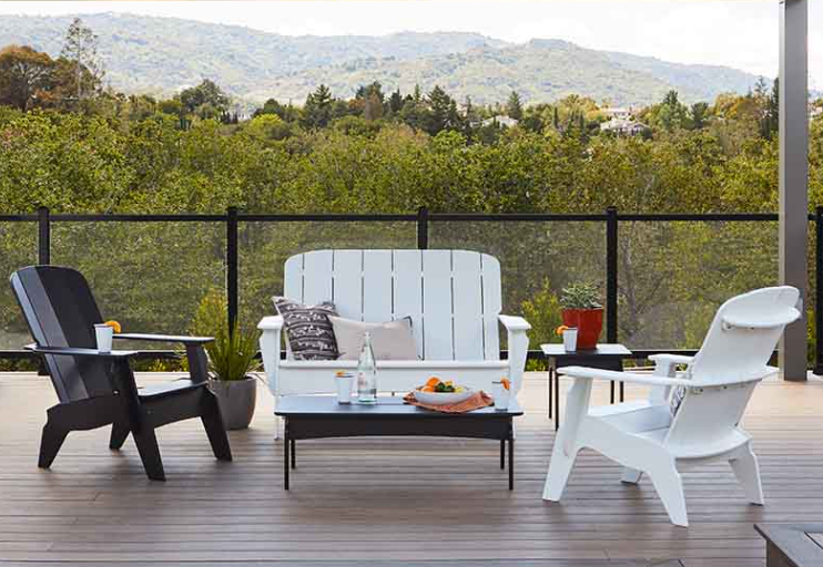A patio features a bright green outdoor sofa, a dark wooden side table with potted plants, and a white, shingled wall.