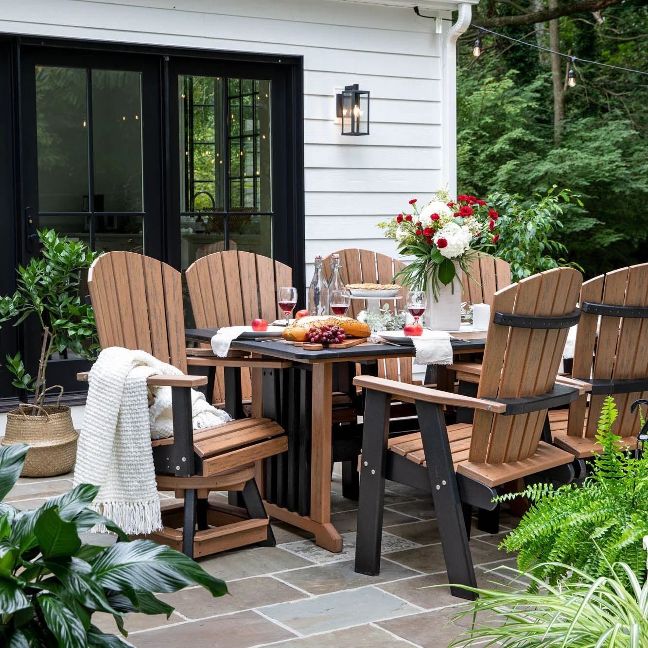 Cozy outdoor patio with wooden chairs, round table, flowers, and food beside a house and garden greenery