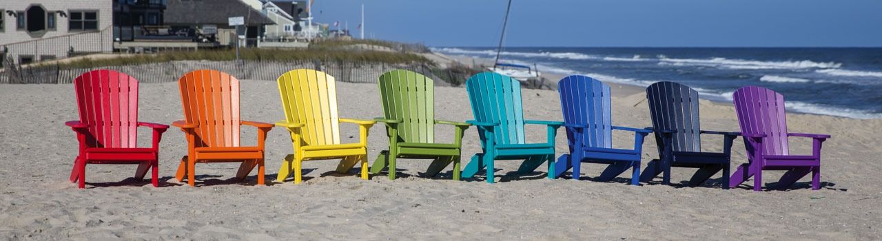 Eight colorful Adirondack chairs lined up on a sandy beach with ocean and houses in the background