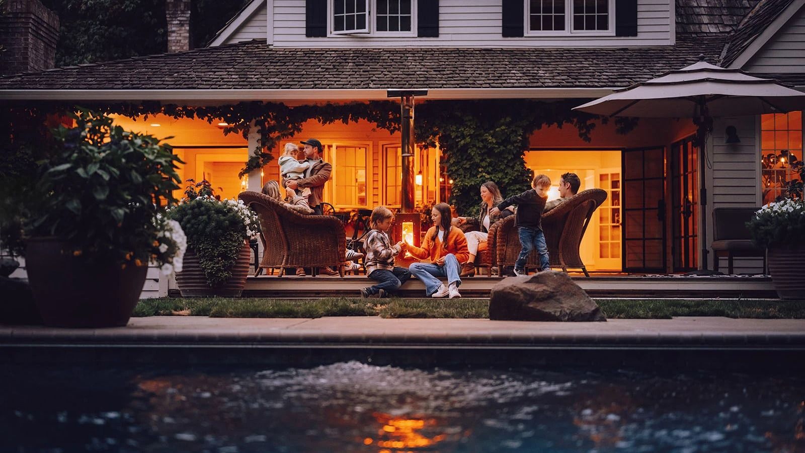 Cozy outdoor patio with wooden chairs, round table, flowers, and food beside a house and garden greenery