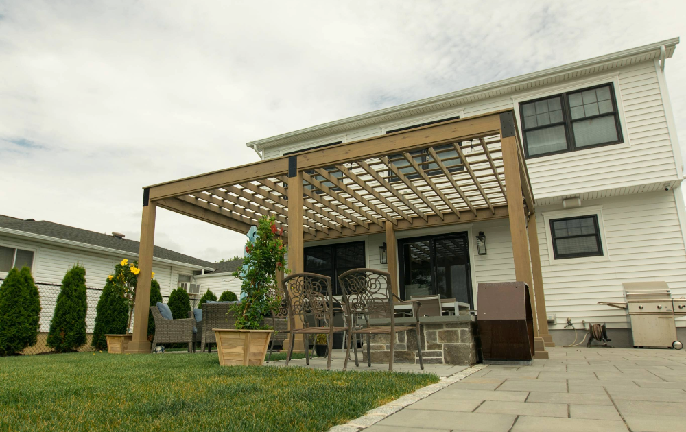 A white pergola with square columns and shutter-style walls stands over a light-colored patio walkway in a bright space.