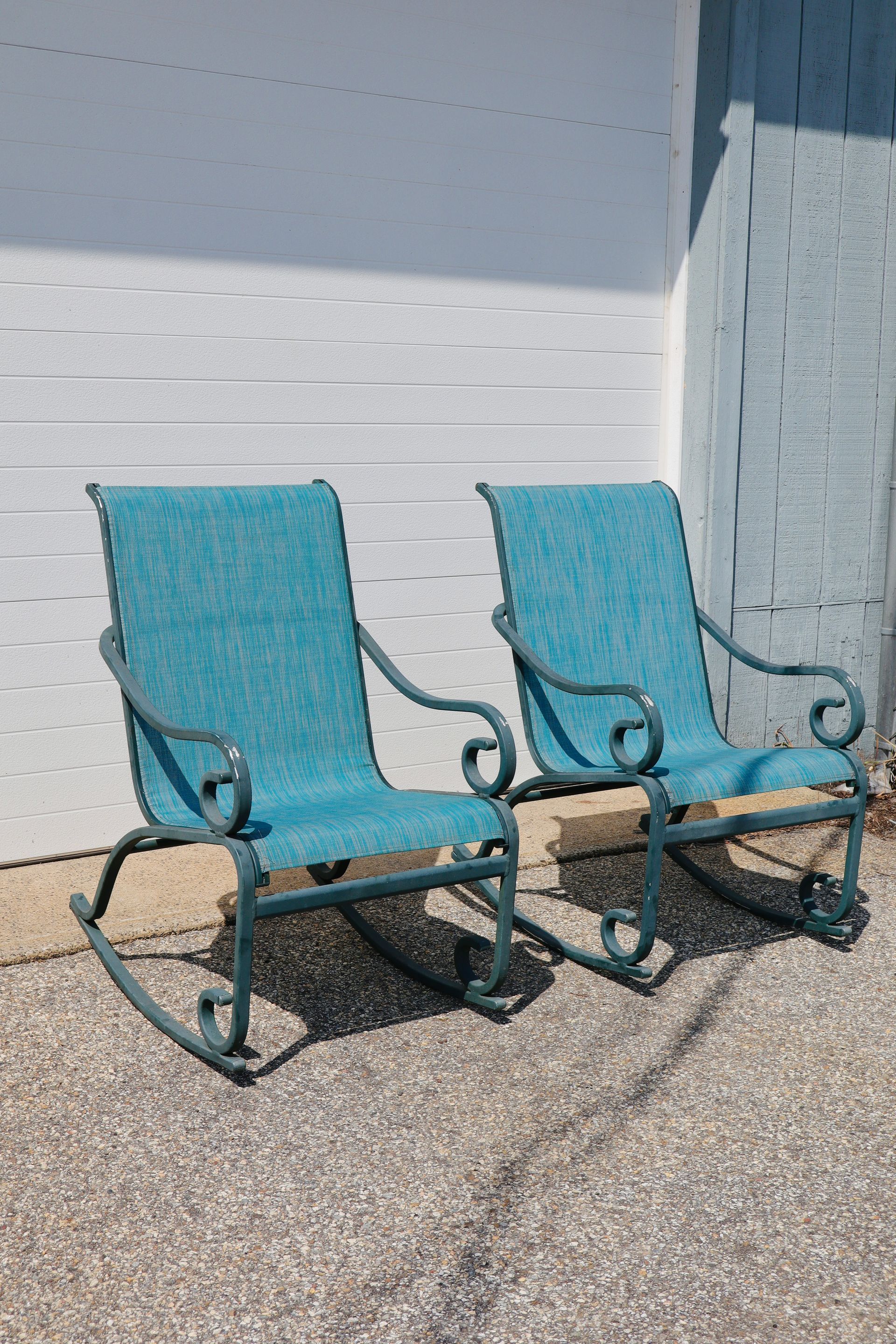 Two turquoise metal patio chairs on gravel beside a light gray wall.