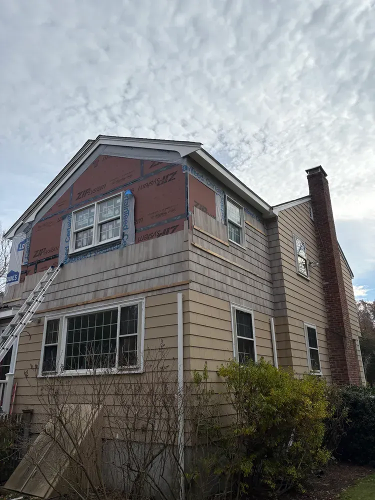 A two-story house exterior under construction with partial siding, exposed house wrap, and a ladder leaning against it.