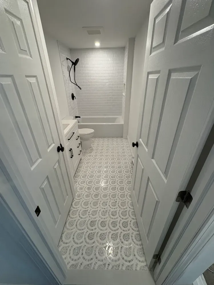 A small, white bathroom featuring patterned grey and white tile flooring, a bathtub, and a pedestal sink.