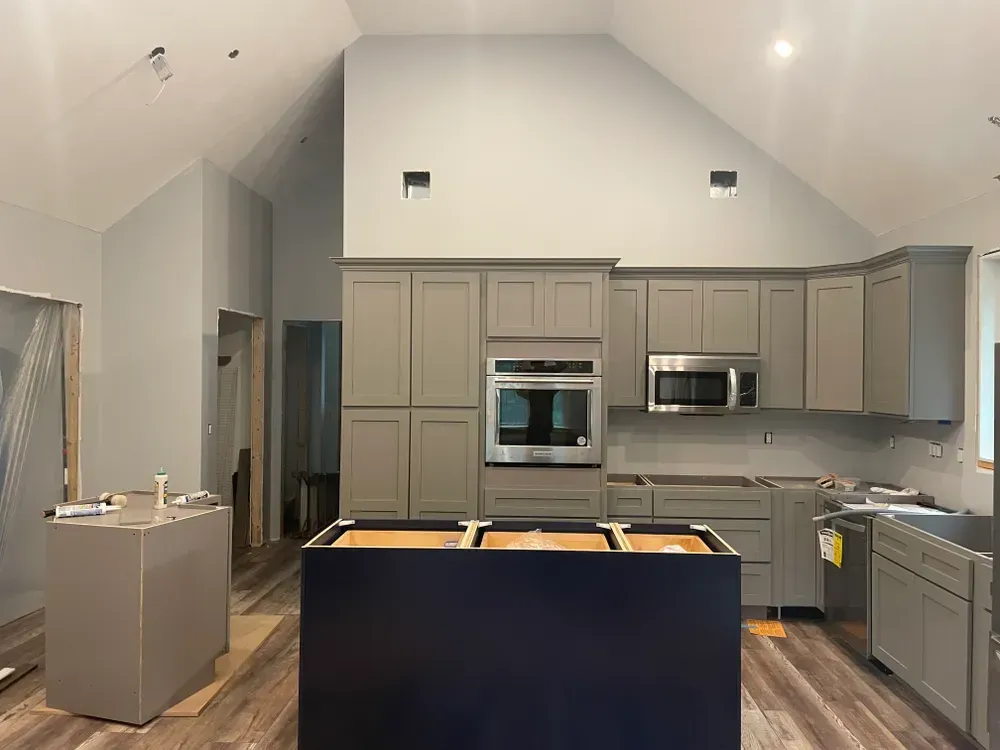 A kitchen under construction with gray cabinets, a blue island base, wood-look flooring, and a high, sloped ceiling.
