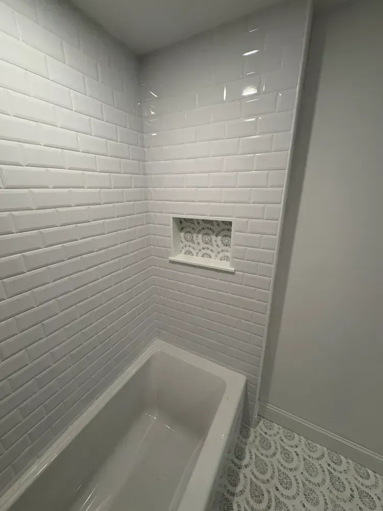 A bathtub with white subway tile walls, featuring a decorative scalloped tile niche and matching floor tiles.