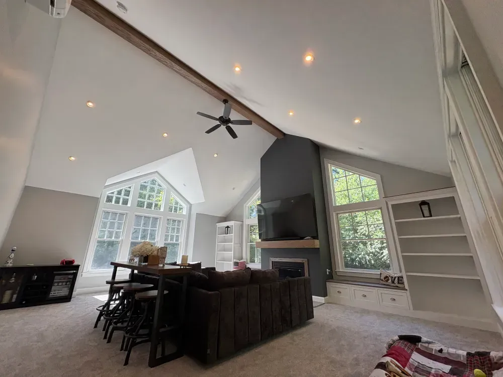 A high-ceiling living room with a large window, dark sofa, bar table, fireplace with a mounted TV, and built-in shelving.