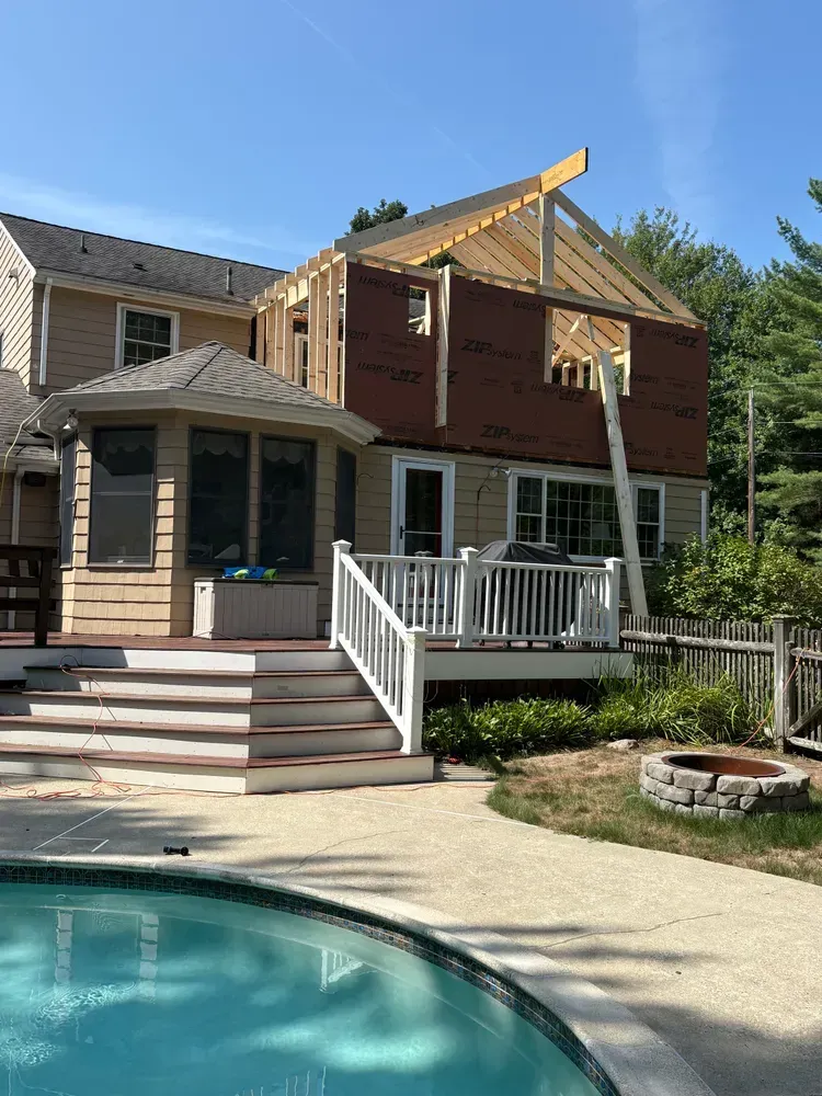 A beige two-story house with a wooden frame addition under construction above a back deck next to a backyard pool.
