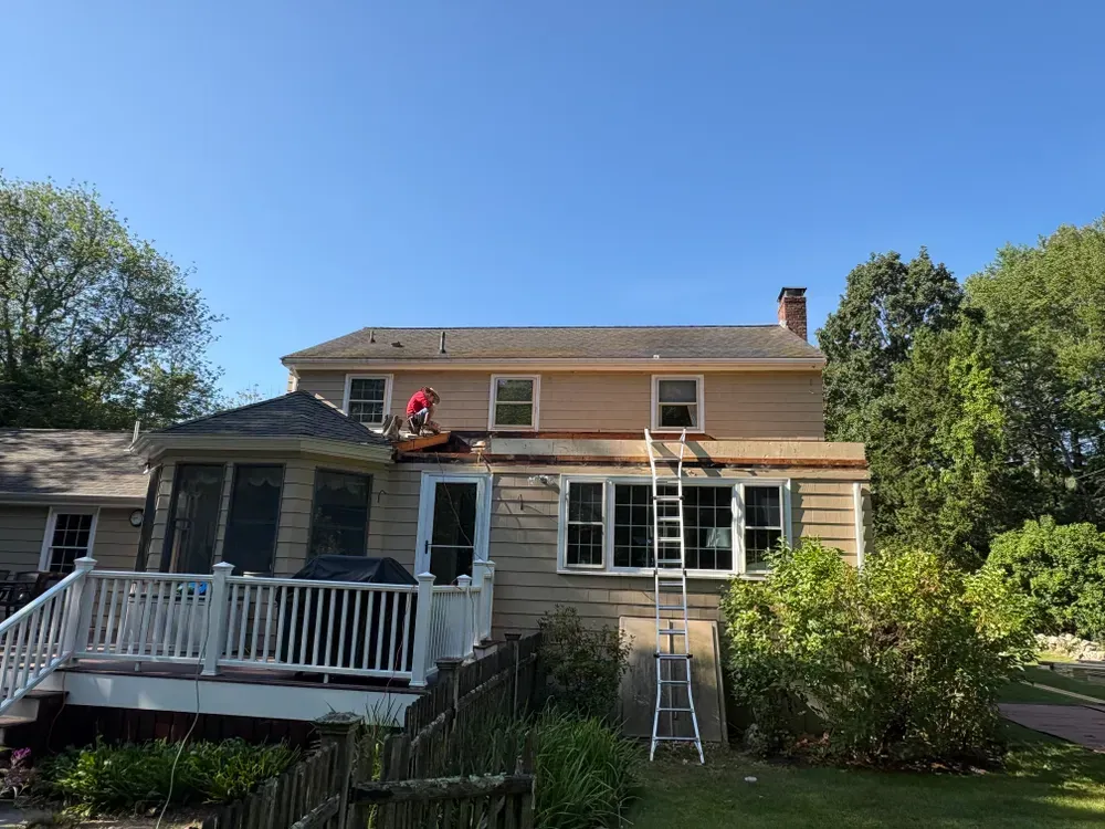 A person in a red shirt works on the roof of a beige two-story house with a white deck, under a clear blue sky.