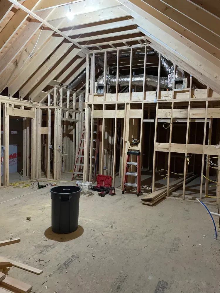 A wide-angle view of a room under construction with exposed wooden wall studs, ceiling rafters, and flexible HVAC ducts.