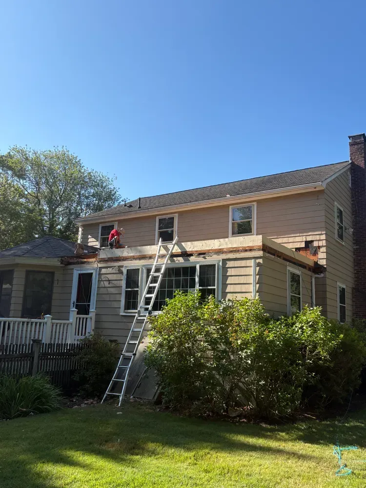 A person on a ladder repairs the roof of a beige two-story house under a clear blue sky.