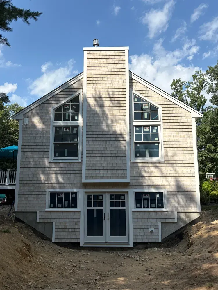 A two-story shingled house exterior with a central chimney, symmetrical windows, and double doors facing a dirt yard.