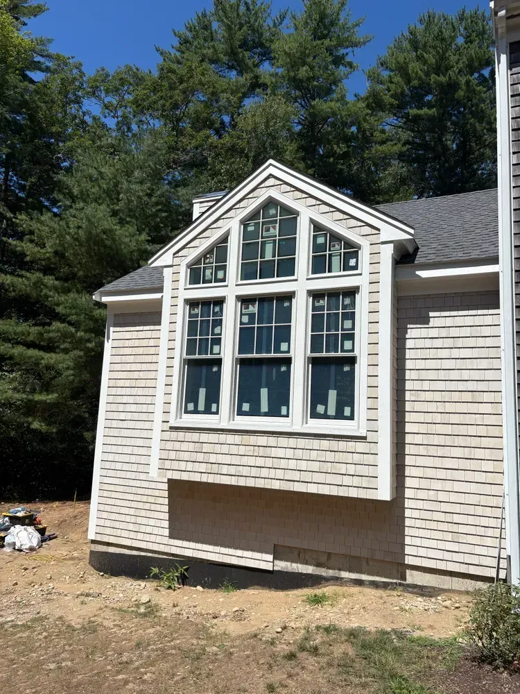 A light-colored house with cedar shingle siding featuring a prominent bay window under a gabled roof set against a forest.