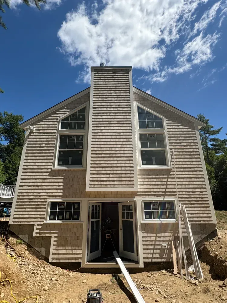 Exterior of a two-story house with beige wood siding, a central chimney, large windows, and open doors under construction.