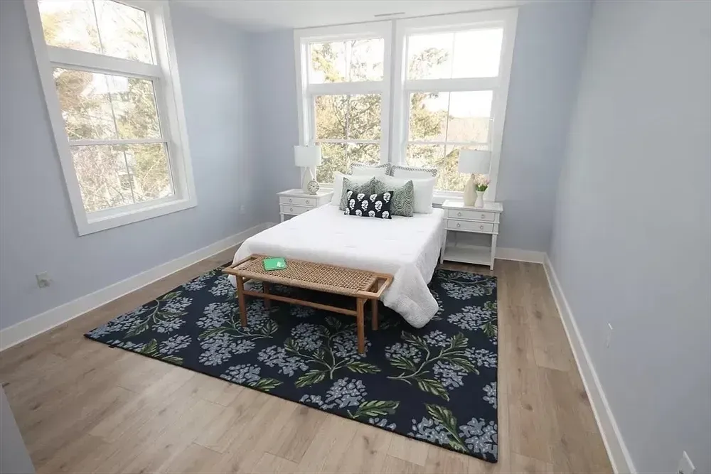 A bedroom with light gray walls, light wood floors, a white bed, a wooden bench, and a dark blue floral area rug.
