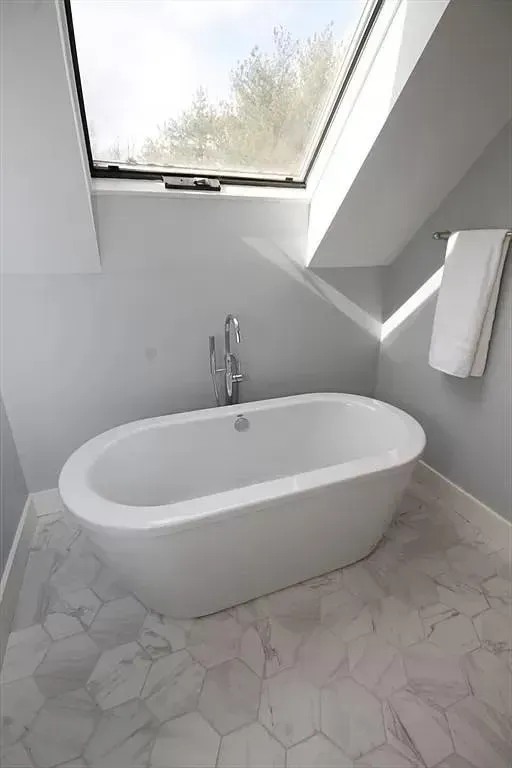 Freestanding white bathtub under a sloped skylight in a bathroom with light grey walls and hexagonal marble floor tiles.