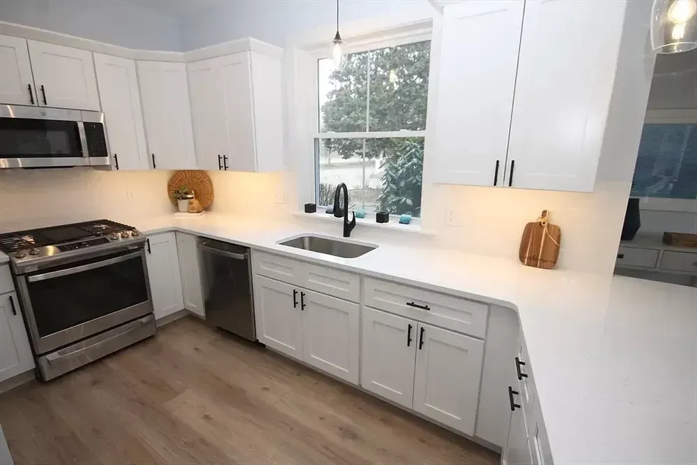 Modern white kitchen with stainless steel appliances, white countertops, dark hardware, and light wood-tone flooring.