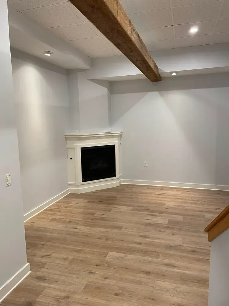 A room with light-colored walls and wood-look flooring features a corner fireplace with a white mantel and a wooden beam.