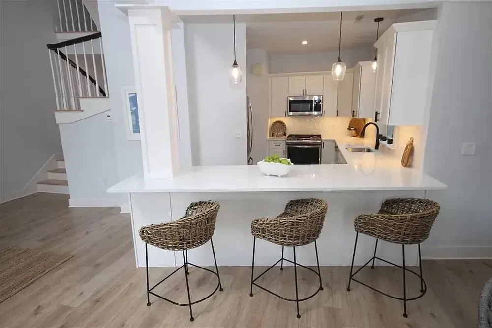 A kitchen with a white quartz breakfast bar, three woven stools, pendant lights, and stainless steel appliances.