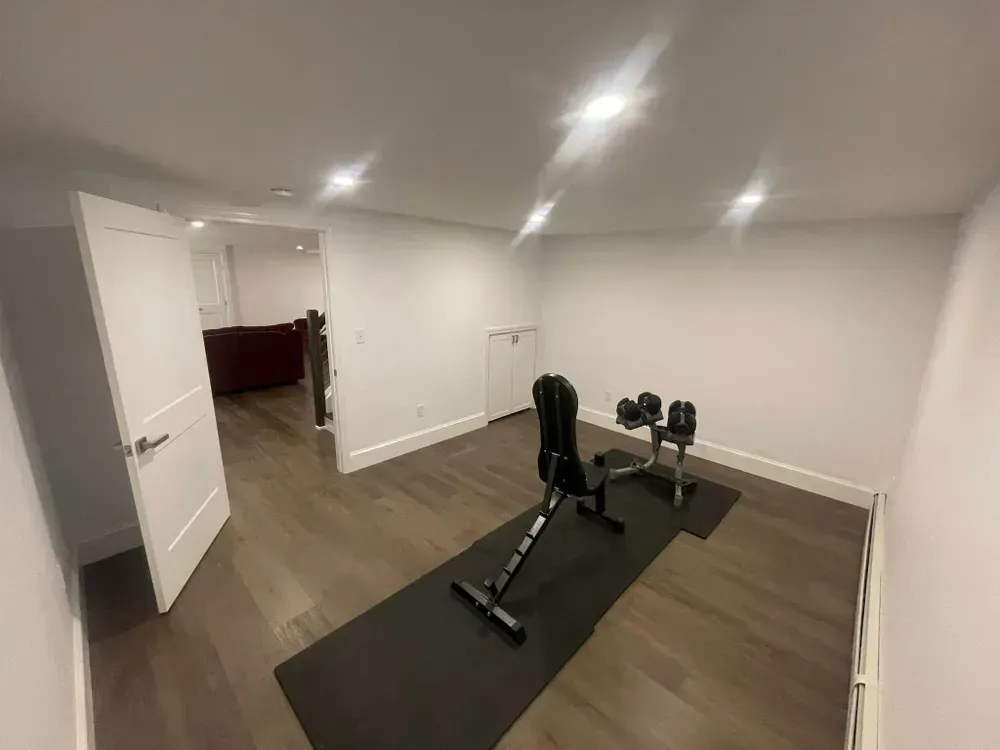 A home gym space with a weight bench and adjustable dumbbells on a black floor mat in a room with white walls.