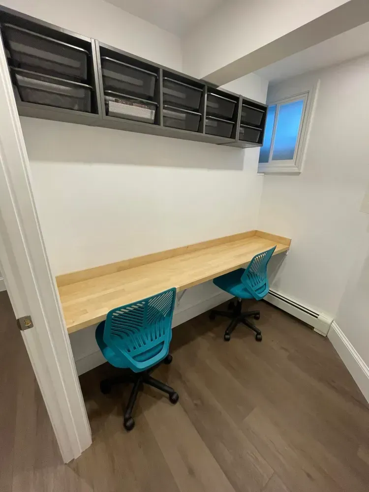 A narrow home office with a long wooden desk, two teal swivel chairs, and overhead storage bins against a white wall.
