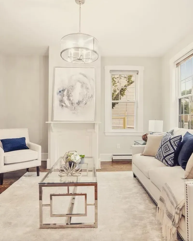A bright, modern living room with a beige sofa, white armchair, glass coffee table, and an abstract painting above a fireplace.
