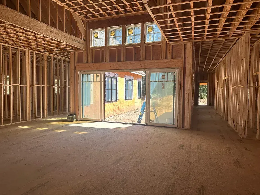 Interior view of a residential building under construction with exposed wooden framing and large glass doors.