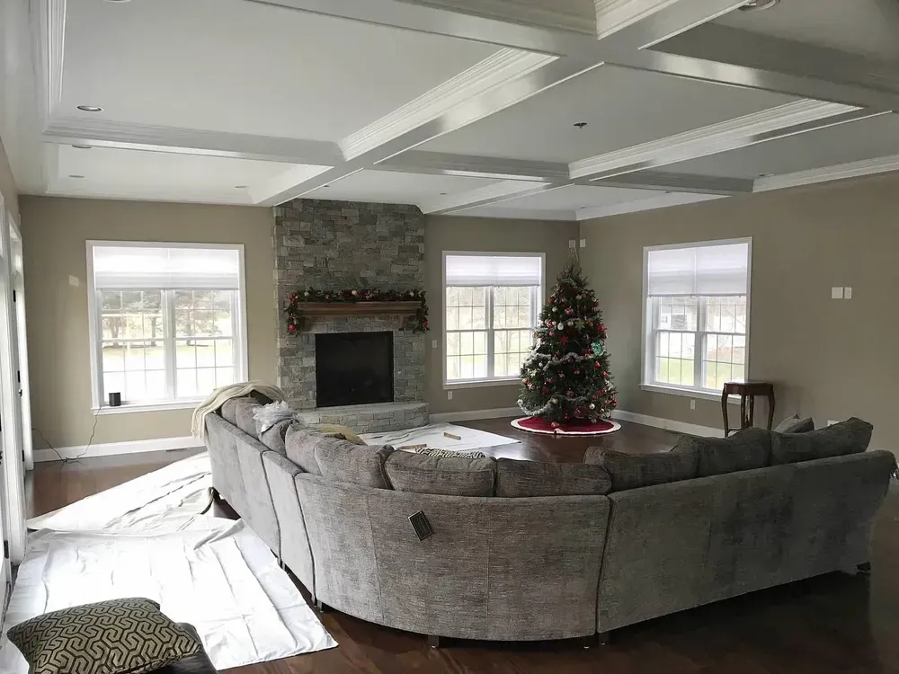 A beige living room features a large grey sectional, a stone fireplace, and a Christmas tree, with coffered ceilings.