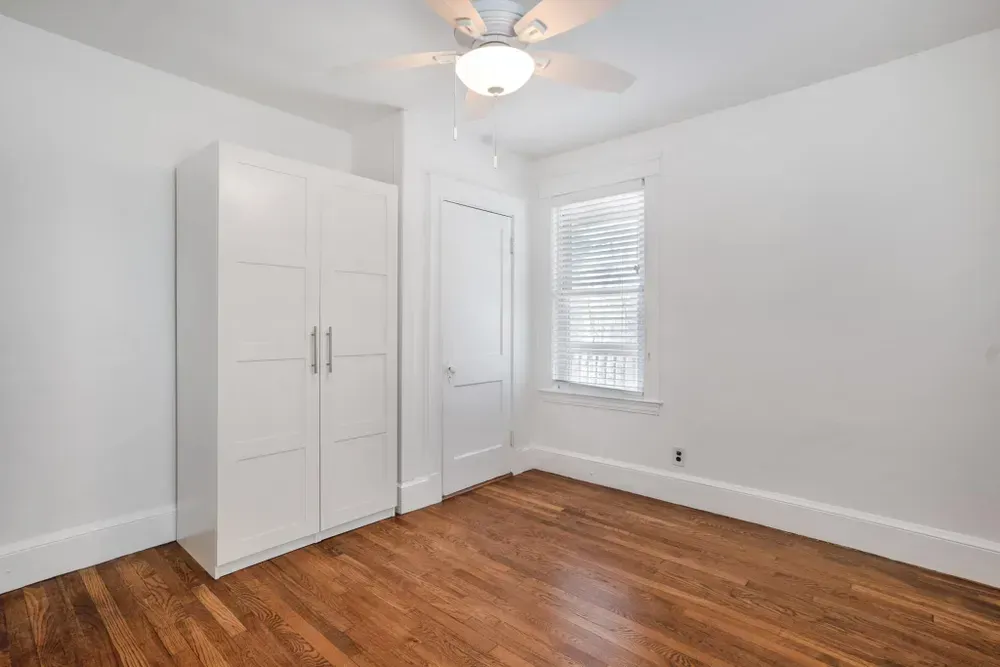 An empty room with white walls, a white wardrobe, a ceiling fan, a window, and wooden floors.