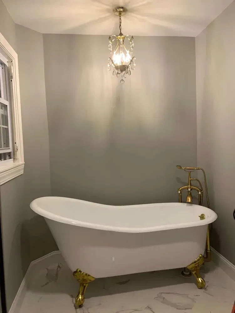 A white clawfoot bathtub with gold hardware sits in a room with grey walls and a crystal chandelier.