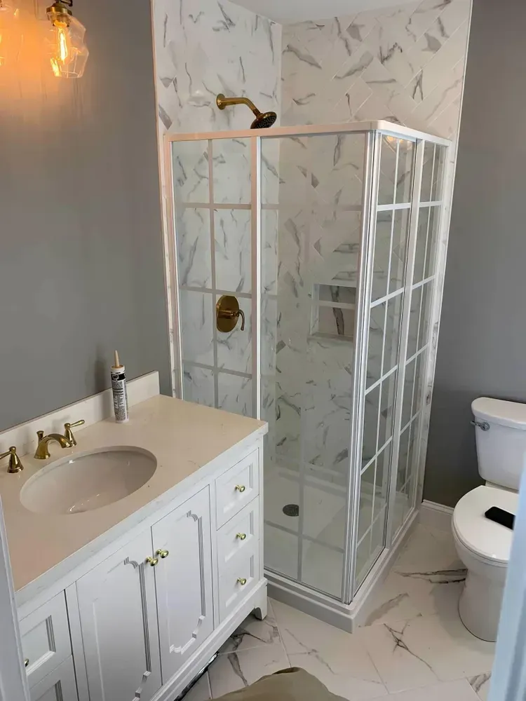 A bathroom featuring a white vanity with a gold faucet, a glass-enclosed shower with marbled tile, and a white toilet.
