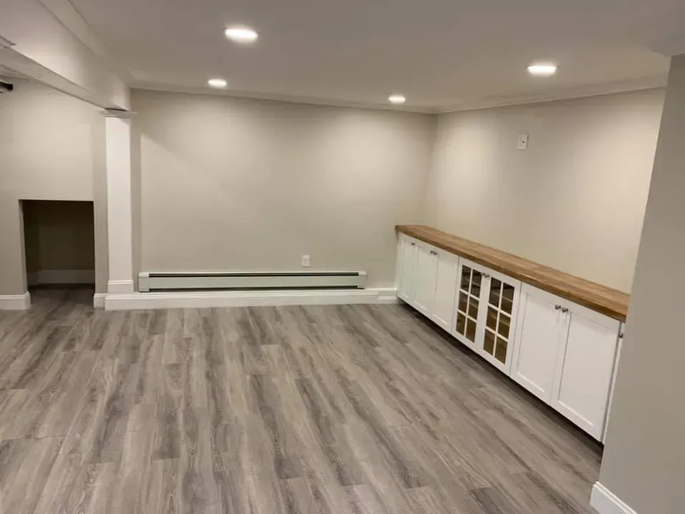 A renovated basement with light gray walls, gray wood-look flooring, recessed lighting, and a long white cabinet console.