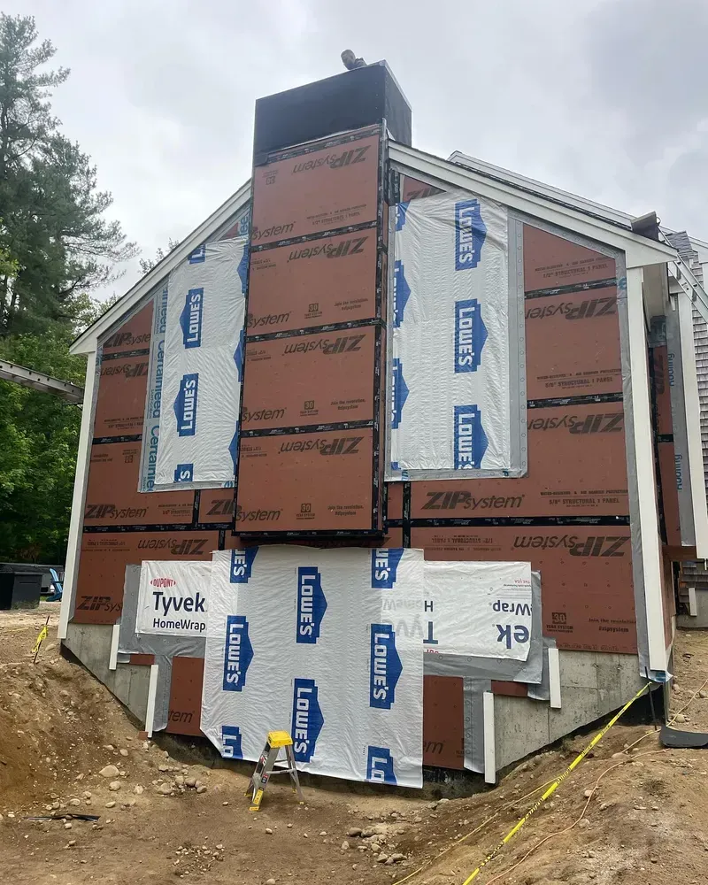 An exterior view of a house under construction, showing framing covered with brown Zip System panels and white vapor barriers.