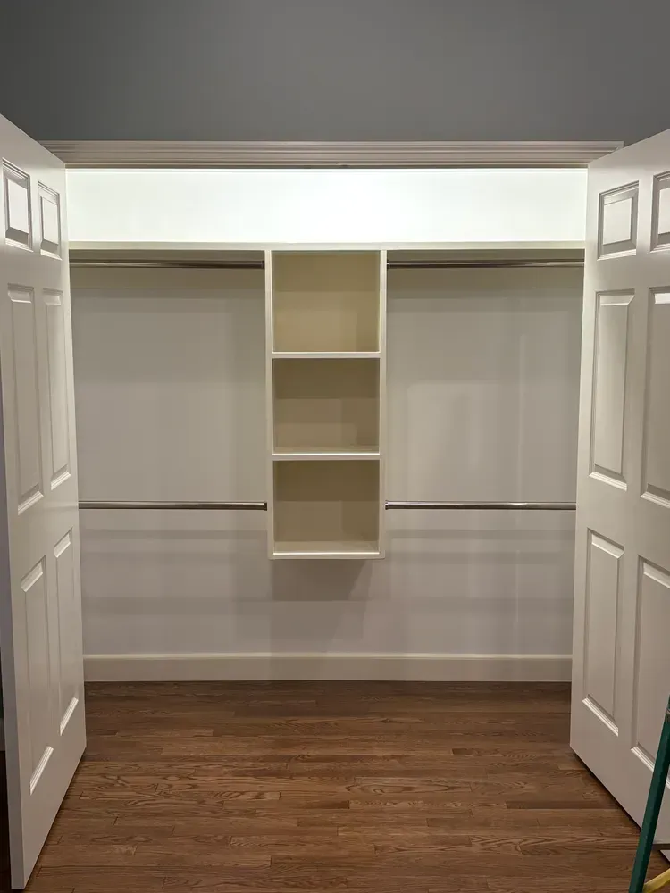 A closet with white doors, hardwood floors, two clothing rods, and a center shelving unit with three open compartments.