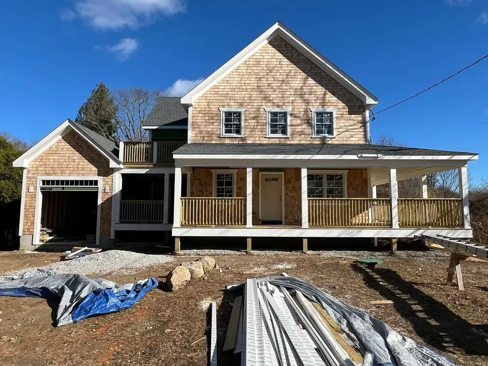 A large, new two-story house under construction with cedar shingles, a covered front porch, and a gravel yard.