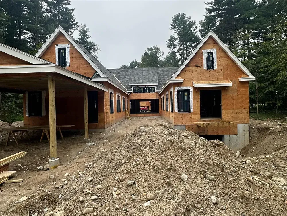 A new house under construction with exposed wood sheathing, a partially built carport, and a dirt mound in the foreground.