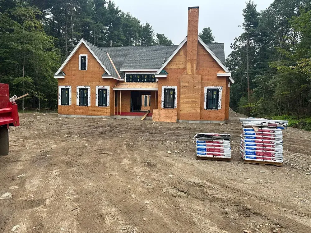 A newly constructed wood-framed house under a cloudy sky, with stacks of building materials in the dirt yard.